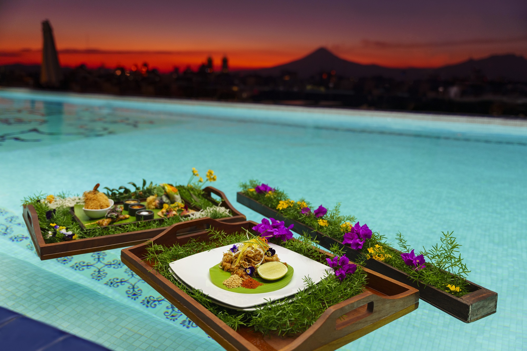Trays with gourmet meals floating in Saffron's pool at sunset, adorned with flowers.